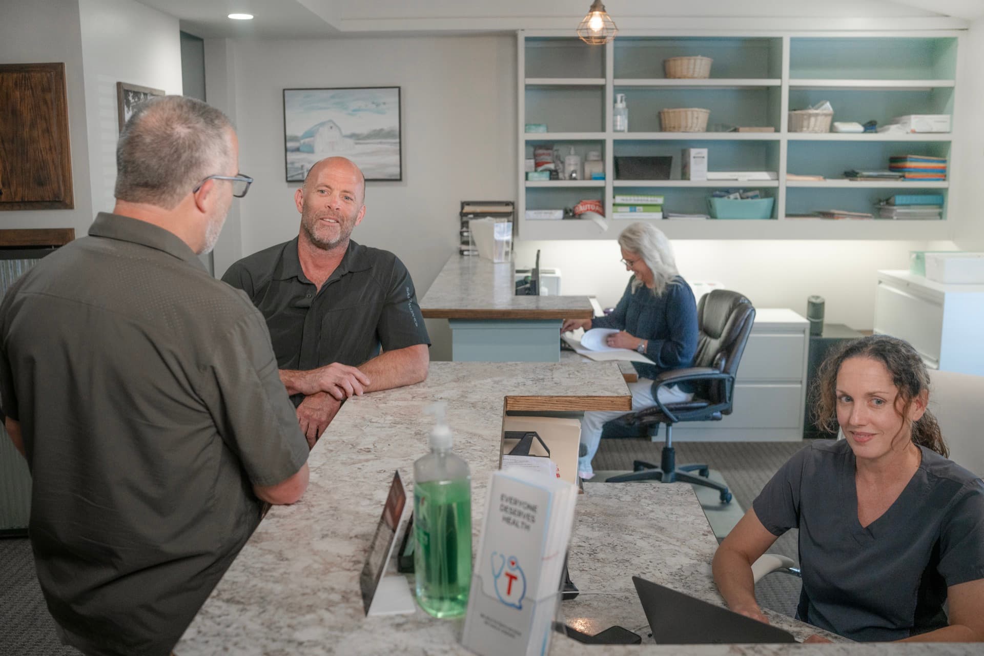 A patient talking with Mark at the front desk while clinic staff work behind it